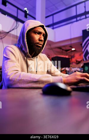 Confused african american criminal using computer for coding illegal ransomware. Concerned young hacker in hood programming malicious software to break into database server Stock Photo