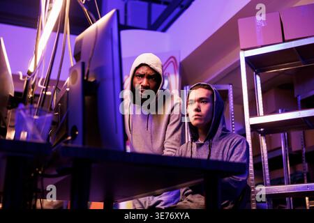 Two concentrated hackers collaborating to crack complex password and break into network system. Young diverse men in hoods working on computer together and coding virus malware Stock Photo