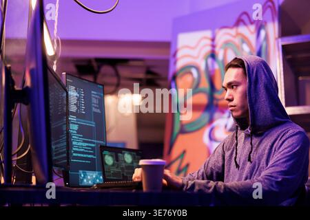 Asian hacker stealing information from server using computer worm. Criminal in hood coding illegal internet virus and breaking into server while drinking coffee to go in dark room Stock Photo