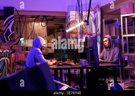 Internet criminals working together to break into database in dark room. African american and asian young men hacking computer system and installing virus malicious software Stock Photo