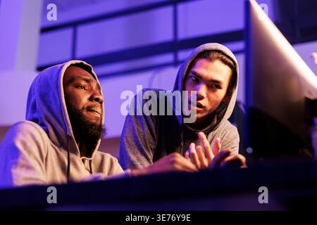 Hackers collaborating while discussing criminal activity and planning cyberattack. African american and asian criminals programming malicious software to break into database Stock Photo
