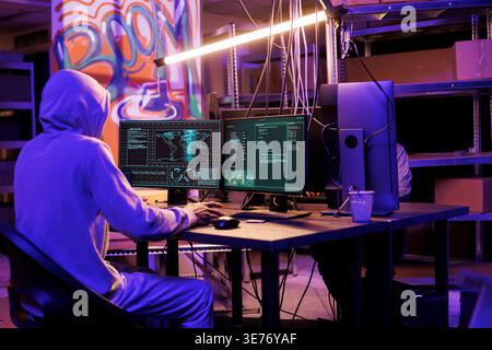 Hacker in hood gathering information using illegal spyware on computer to break into server. Criminal doing web tracking with malicious software and checking data on screen Stock Photo