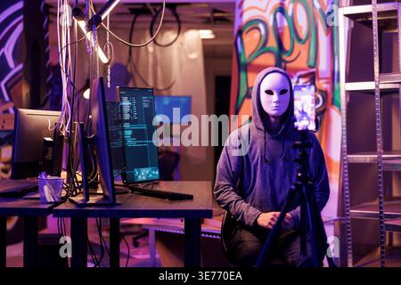 Anonymous hacker recording threat video with ransom message on smartphone to steal privacy information. Internet criminal calling victim using mobile phone in abandoned warehouse Stock Photo
