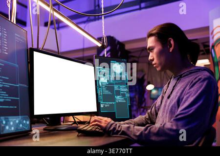 Hacker spreading malware and doing illegal activities on computer with white screen. Young asian criminal breaking law while programming malicious malware in abandoned warehouse Stock Photo