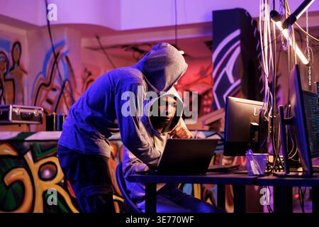 Criminals hacking on laptop together and planning ddos attack on website. Two hackers in hoods checking malcious software code on portable computer in abandoned warehouse Stock Photo
