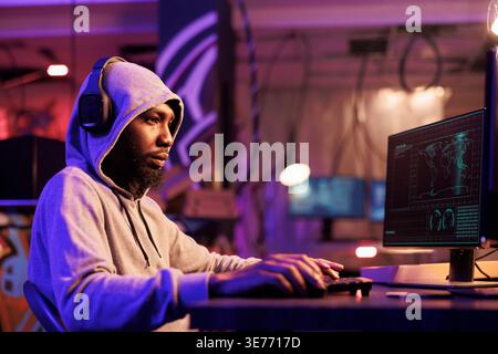 Hacker in headphones writing malware code and typing on keyboard. Internet criminal in hood listening to music in earphones and breaking into database server to breach data Stock Photo