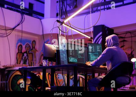 Hacker developing internet virus for data breaching and hacking server in dark warehouse. Man in hood running malware code for password cracking on computer screen in room at night Stock Photo