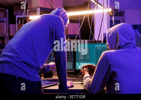Two hackers running malicious software on computer screen to breach secure data. Criminals group wearing hoodies while breaking law and working together in abandoned warehouse Stock Photo