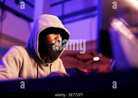 African american hacker breaking into database to steal sensitive information. Man in hood accessing computer system illegally and coding malicious software for cyberattack in hideout Stock Photo