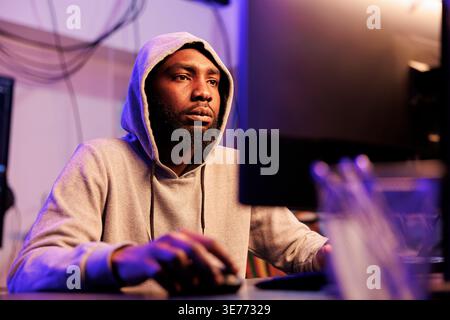African american hacker coding virus malicious software on computer and breaking law. Criminal in hood using stolen password to breach company server and commit online fraud Stock Photo