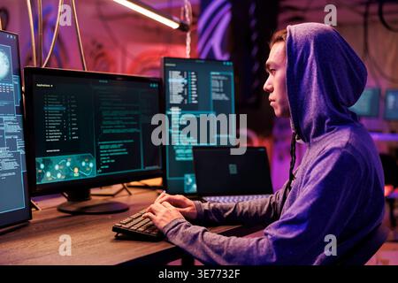 Asian hacker typing on keyboard and writing spyware code on computer. Internet criminal in hood hacking online server and planning phishing cyberattack in room at night time Stock Photo