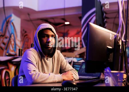 African american hacker doing illegal activities on computer and working late portrait. Criminal in hood breaking website with ddos attack and looking at camera in abandoned warehouse Stock Photo