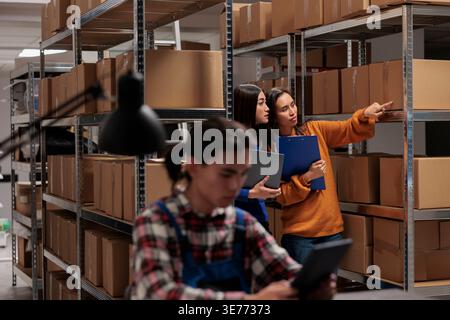 Storehouse asian women employees checking package packing quality in postal storage room. Storehouse shipment operator showing goods in stock on shelf to logistics manager Stock Photo