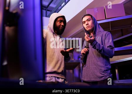 Hackers discussing ddos attack technique to break website while standing near computer. African american and asian criminals planning to breach cybersecurity system and steal confidential data Stock Photo
