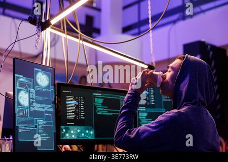 Hacker drinking coffee and attempting malware attack on web server. Young man in hood doing criminal activity and breaking law while sipping beverage to go and breaching data Stock Photo