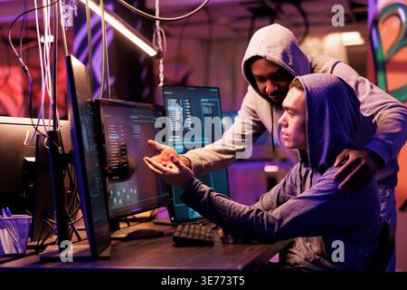Two hackers collaborating while breaching data and coding malicious software on computer. Diverse criminals working together while programming ransomware in abandoned warehouse Stock Photo
