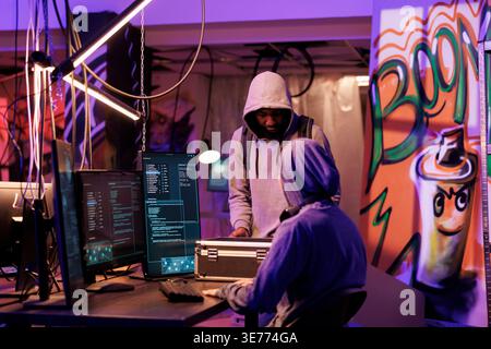 Hacker in hood showing black box with hacking technology to break into server database. Young criminal opening case with equipment for online cyberattack in abandoned warehouse Stock Photo