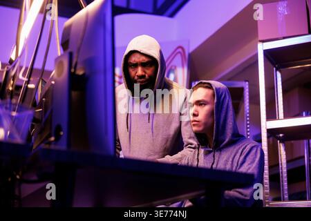 Hackers identifying vulnerabilities in system to break into database and steal data. Asian and african american criminals in hoods hacking online server and cracking password on computer Stock Photo