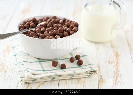 Chocolate corn balls in a ceramic bowl on a white wooden table, selective focus Making a healthy breakfast cereal using chocolate corn balls and milk Stock Photo