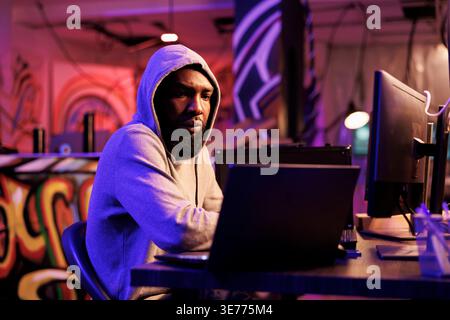 African american hacker coding computer virus to attack database. Young developer in hood cracking password and using spyware to break into online server and steal information Stock Photo