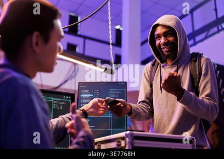 Smiling cheerful illegal malware developer holding ssd with computer virus. Happy hacker giving hard drive with malicious software to crime partner to steal information from database Stock Photo