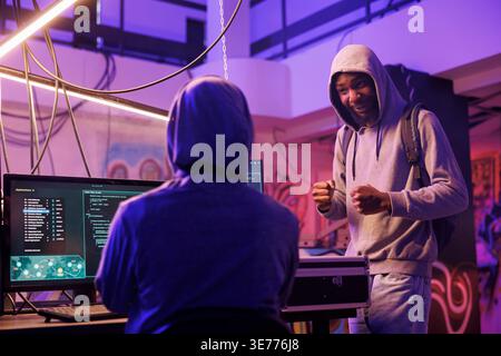 Cheerful internet scammers discussing phishing scam and planning online fraud. Smiling african american hacker explaining data breach idea to criminal in abandoned warehouse Stock Photo