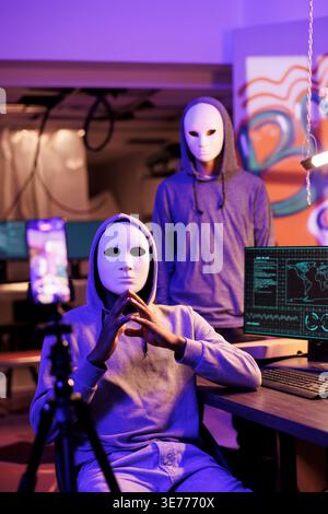 Scammers in anonymous masks streaming threat online and demand victim for payment. Hackers with hidden identity filming ransom message on mobile phone in abandoned warehouse Stock Photo