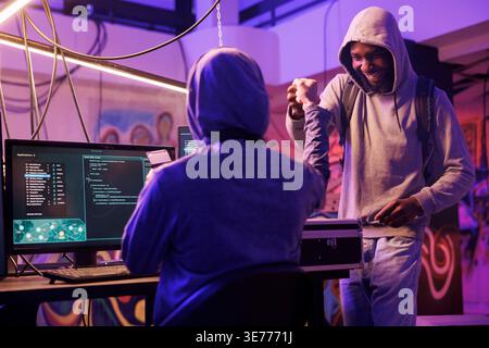 Cheerful african american hacker giving fist bump to crime partner while working together in abandoned warehouse. Smiling men in hoods sharing friendly gesture after successful database breaking Stock Photo