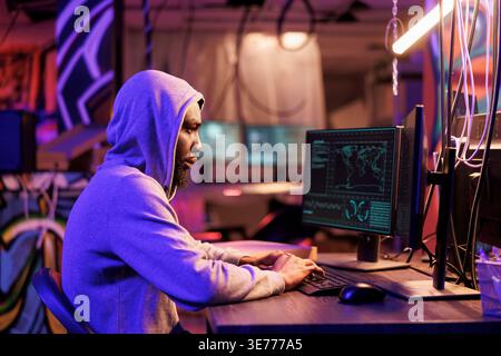 African american hacker coding spyware in abandoned warehouse. Man in hood using computer for breaching data, cracking password and getting access to server database at night Stock Photo