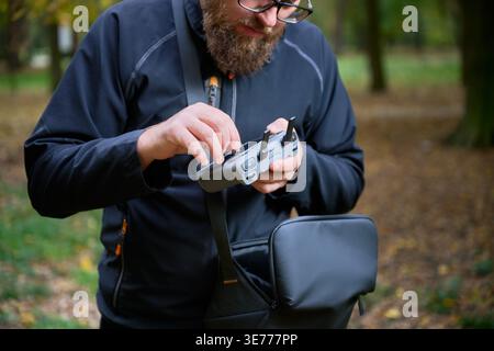 Man adjusts drone controller and checks settings before flight. Bearded man preparing remote control in a park, setting up equipment for aerial photography and filming Stock Photo