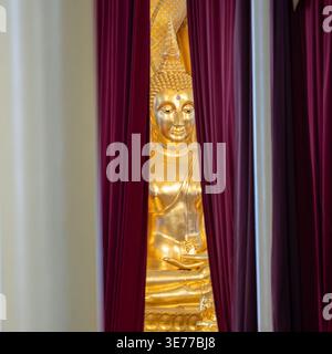 A golden Buddha statue appears between deep red curtains inside the temple. Draped fabric frames the shrine view. Stock Photo