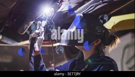 Welding female tech inspecting car underside in garage, using torch, helmet, gloves, blue overlays Stock Photo