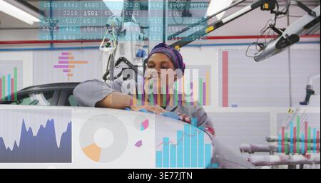 Wiping tech in gray coverall, scarf, leaning on white car in workshop, blue cloth, digital charts Stock Photo