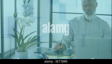 Senior African American man in shirt using laptop charts, drawing on tablet with stylus, copy space Stock Photo