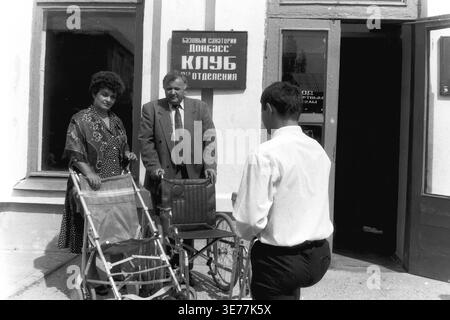 Slovyansk Resort, 1997. A poignant black and white archival photograph taken outside the club building of the Donbas sanatorium. A middle-aged man and woman stand solemnly next to two empty manual wheelchairs, while a younger man faces them, partially seen from the back. This powerful vintage image documents the essential medical and rehabilitative social infrastructure of the famous local mud-bath resort, capturing a moment of personal significance and healthcare accessibility in a peaceful Donbas. Stock Photo
