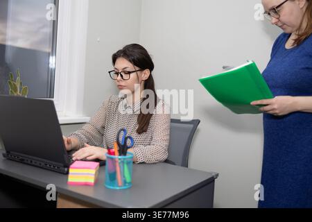 One woman types on a laptop at her desk. Another woman stands near her with a folder. The setting is an office with a window and some decorations. Stock Photo