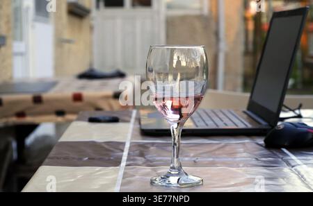Outdoor workspace scene with a glass of light red wine next to an open laptop and mouse on a checkered tablecloth, in soft daylight with blurred backg Stock Photo