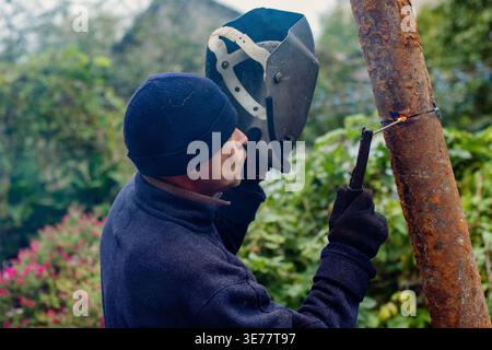 This image captures a focused male worker outdoors, engaged in arc welding a rusty metal pipe. With his dark blue beanie and protective gloves, he ski Stock Photo