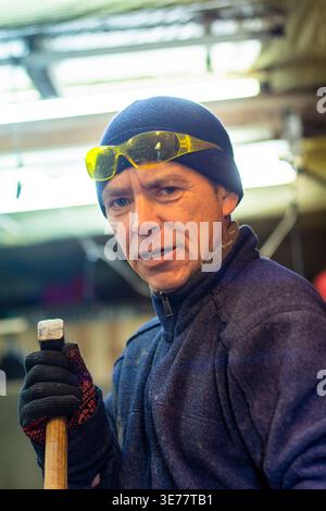 A portrait of a focused middle-aged male worker in an industrial workshop environment, wearing safety glasses and a blue jacket while holding a heavy Stock Photo