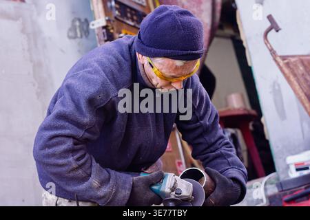 A professional worker in protective gear carefully cuts a steel pipe using a handheld angle grinder in a workshop environment. Stock Photo