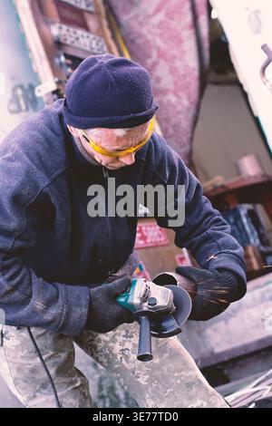 A focused male worker in safety goggles and gloves uses a powerful angle grinder to cut through a metal pipe, creating a stream of bright sparks in a Stock Photo