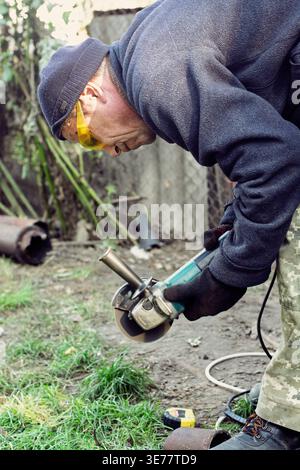 A man in protective gear uses a power angle grinder to cut a metal pipe in an outdoor yard, focusing on precision and home repair tasks. Stock Photo