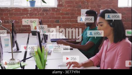 Typing duo wearing pink blouse and green tee in open-plan office with headset, screens and pings Stock Photo