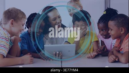 Smiling teacher in light blouse guiding seven kids at class table, holding tablet with blue overlay Stock Photo