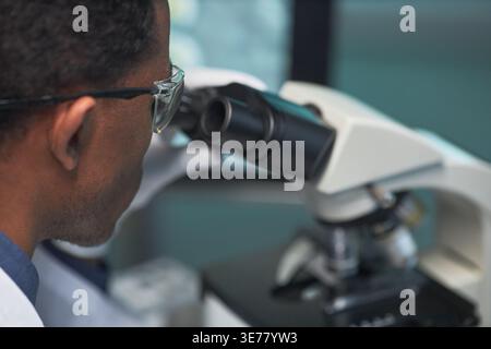 Close up of Black male scientist examining sample through microscope in laboratory, supporting medical research and diagnostics. Ideal for healthcare innovation or biotech marketing Stock Photo
