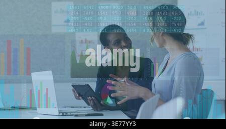 Discussing data, two women wearing blouse blazer gesturing at meeting with laptop, tablet overlays Stock Photo