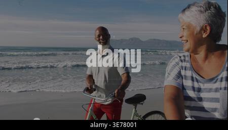 Chatting man holding bike and woman riding bike on sandy beach, wearing polo and striped shirts Stock Photo