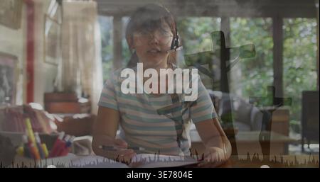 Studying girl wearing striped tee, headset, specs, writing in book at home with three-cross overlay Stock Photo