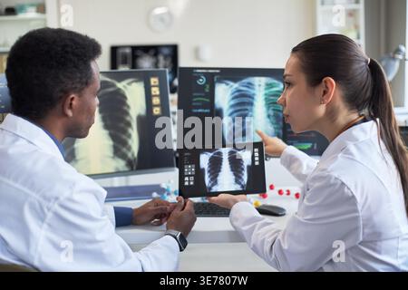 Medium shot of Black male doctor and Asian female doctor reviewing chest x rays on monitors and tablet, discussing diagnosis. Ideal for healthcare teamwork, radiology, telemedicine marketing Stock Photo