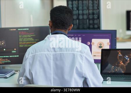 African American male scientist analyzing genomic data on multiple screens in laboratory, supporting biotechnology research and precision medicine marketing, rear view Stock Photo